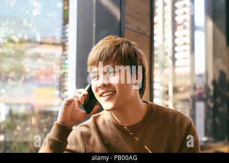 Closeup portrait of smiling Young Asian man talking on mobile phone et assis à table vide intérieur en arrière-plan flou cafe Banque D'Images