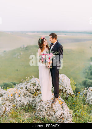 Couple cherche dans les yeux de l'autre debout sur le rocher au fond de la montagne verte. Banque D'Images