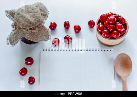 Les cerises mûres rouges avec pot de confiture, cuillère en bois et portable sur fond blanc. Mise à plat. Concept alimentaire. Banque D'Images