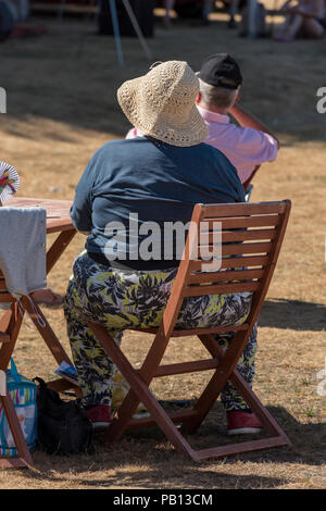 Grand, gras obèse femme assise sur une chaise portant un chapeau de soleil sur une journée chaude. Banque D'Images