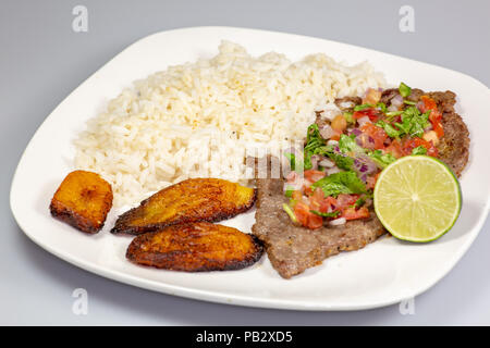 Un steak couverts de pico de gallo entouré par la banane plantain et le riz blanc sur une plaque blanche. Cuisine cubaine. Banque D'Images