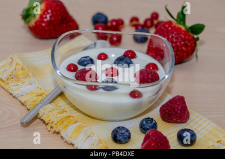 Framboises, bleuets, fraises et des mûres avec du yogourt dans un bol en verre sur une serviette jaune sur une table en bois Banque D'Images