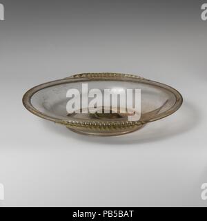Plat en verre. Culture : Roman. Dimensions : diam. : 8 3/16 in. (20,8 cm). Date : fin 1er-début du 3e siècle de notre ère. Pâle translucide vert gris fortement teinté de violet ; poignées de même couleur. Large jante tubulaire, faite en repliant vers l'extérieur et vers le bas, presque à l'horizontale en forme de S ; l'épaule d'un côté à l'organisme ; le joint torique de la base tubulaire, faite par pliage ; bas presque plat mais avec un dôme épais au centre avec des traces de grand circulaire pontil marque ; deux poignées le long du bord de la jante traîné de droite à gauche. Sur les poignées, vertical outillage pour faire une série d'une alternance de crêtes et de gorges. Intacts ; quelques piqûre et deux grandes Banque D'Images