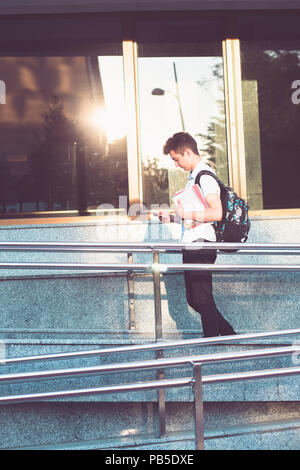 L'aide aux étudiants titulaires d'un téléphone mobile et ordinateur portable de transporter un sac à dos marche à l'avant du bâtiment de l'université. Young boy wearing blue shirt et da Banque D'Images