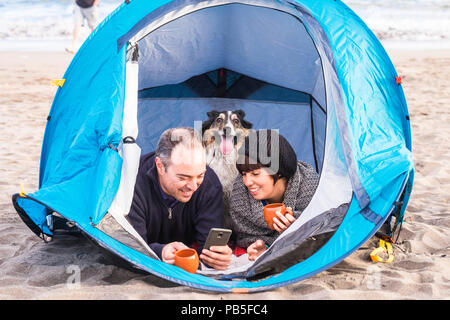 Couple le téléphone intelligent et avoir du plaisir à l'intérieur d'une tente dans un camping sur la plage chien border collie derrière eux regardant la caméra. Banque D'Images