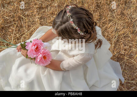 Petite fille vêtue de première communion avec des fleurs à la main assis sur la paille Banque D'Images