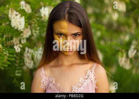 Portrait en gros plan d'une belle jeune fille en blanc autour de l'acacia au printemps la moitié face golden make-up Banque D'Images
