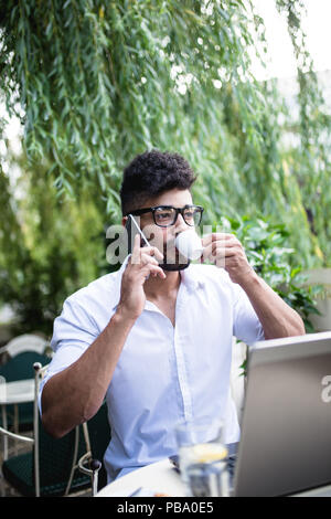 Les jeunes afro-américains attractive businessman sitting in cafe bar faire un certain travail sur ordinateur portable et prendre sur téléphone mobile. Banque D'Images