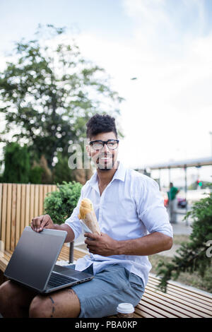 Jeune homme beau avec des lunettes assis sur rue avec ordinateur portable et eating sandwich. Concept d'entreprise. Banque D'Images