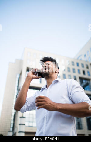 Beau jeune homme afro-américain debout devant d'énormes capacités d'affaires moderne, smiling, holding coffee pour aller et parler sur téléphone mobile. Banque D'Images