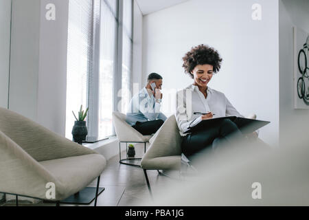 Smiling african woman sitting on office hall travaille sur un rapport d'affaires avec collègue masculin parlant au téléphone en arrière-plan. Banque D'Images