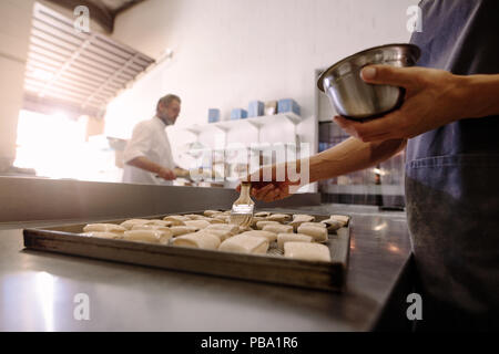 Mains de femme baker avec pinceau beurre frottis sur les rouleaux placés sur la plaque de cuisson en boulangerie. Baker lubrification de la surface supérieure de rouleaux avant leur entrée dans Banque D'Images