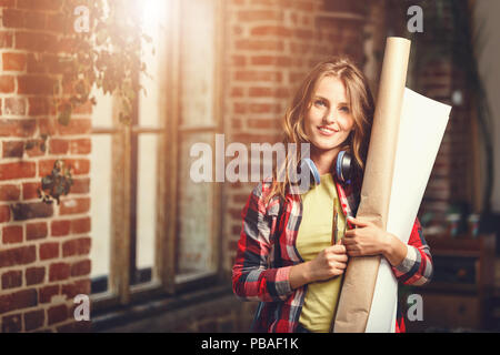 Beau jeune femme cheveux long-architect holding paper rolls et souriant près de la fenêtre de la mezzanine bureau moderne Banque D'Images