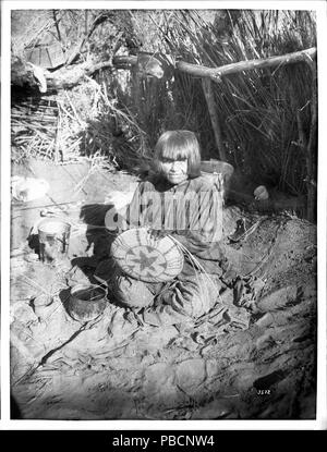 . Anglais : Portrait d'une femme indienne Apache vannier, ca.1900 portrait photographique d'une femme indienne Apache vannier, ca.1900. Elle est assise sur un chiffon sur le sol avec un panier partiellement finis avec une étoile sur le bas dans ses mains sur ses genoux. Plusieurs seaux et pots de céramique s'asseoir autour d'elle. Elle porte une robe à carreaux sur une robe en pointillés. Ses cheveux sont coupés au-dessus de ses épaules et a bangs. Derrière elle est une barrière en bois. Derrière la barrière de hautes herbes se développe. Une boîte en bois est assis sur une branche d'arbre derrière elle à gauche. Numéro d'appel : SHC-3572 Photographe : Pierce, C.C. (Cha Banque D'Images