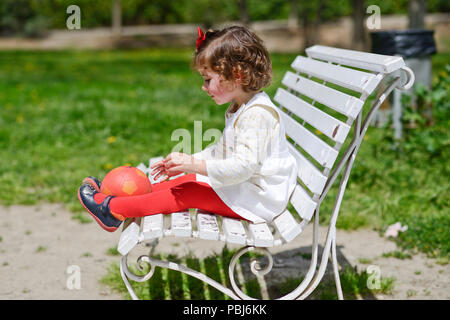 Adorable petite fille jouant avec une balle assise sur un banc de parc Banque D'Images
