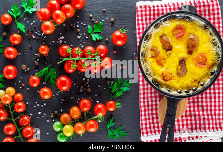 Omelette au plat avec des saucisses et des oeufs de poule dans un pot en fonte ronde et mûrs tomates cerises rouges sur fond noir, vue du dessus Banque D'Images