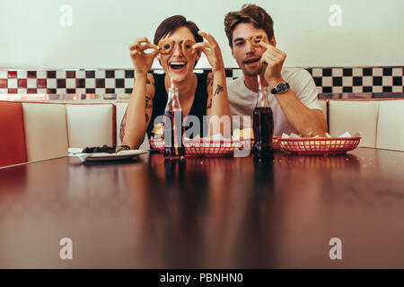 Couple assis dans un restaurant et s'amuser holding biscuit bagues à leur yeux. Smiling man and woman at a diner assis avec de la nourriture et de la SOF Banque D'Images