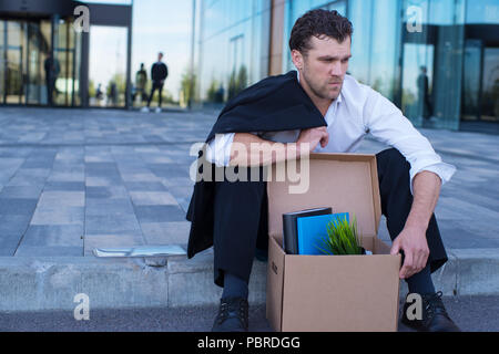 Tiré business man sitting frustré et contrarié dans la rue près de l'immeuble de bureaux de fort de ses affaires. Il a perdu travail Banque D'Images