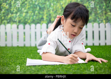 Petite fille asiatique chinois agenouillé sur l'herbe et à faire leurs devoirs à l'extérieur parc Banque D'Images