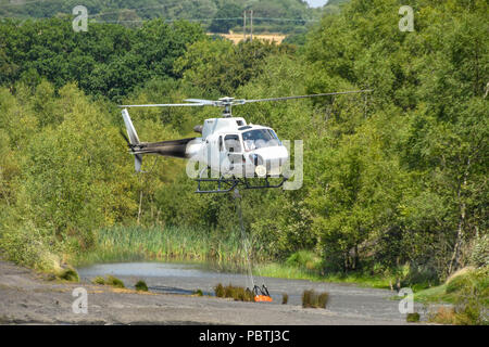 Un hélicoptère en vol stationnaire au-dessus d'un étang pour remplir un grand réservoir d'eau avant de s'envoler pour laisser tomber l'eau sur le site d'un grand feu d'herbe. Banque D'Images