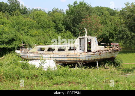 Vieux bateau en bois délabré abandonné avec des fenêtres cassées, portes, panneaux en bois et des pièces de métal rouillé, à gauche dans la cour entre l'herbe non coupée et fores Banque D'Images