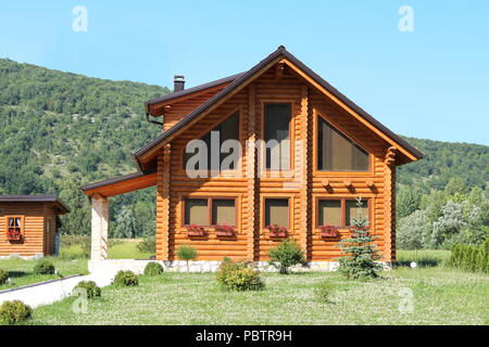 Cabane en bois nouvellement construit maison avec de grandes fenêtres et l'entrée en pierre entouré d'herbe non coupée plein de petites fleurs et forêt avec bleu clair Banque D'Images