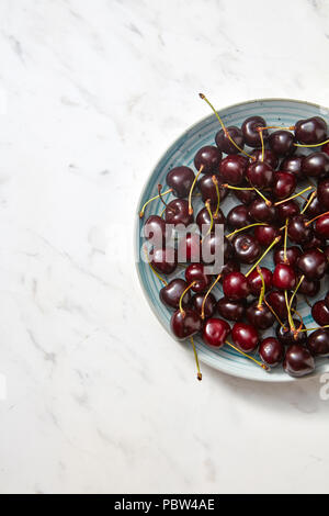Un tas de douces mûres de cerise rouge foncé dans une plaque en céramique bleu sur un fond noir en gris. Mise à plat, demi-plat. Banque D'Images