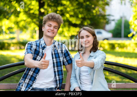 Un gars avec une fille en parc d'été dans la nature. Ils s'asseoir sur un banc en ville. Avec un geste de main montre un pouce levé, comme. Les émotions de bonheur et de plaisir. Concept de succès dans l'atteinte des résultats. Banque D'Images