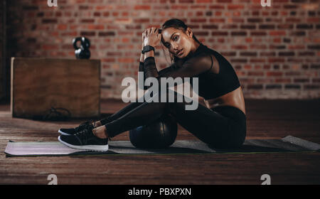 Portrait of tired woman sitting on exercise mat avec ballon sur le côté et à la recherche à huis clos, à la salle de sport. Fit woman se reposant après une formation polyvalente à Banque D'Images
