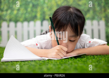 Petite fille asiatique chinois agenouillé sur l'herbe et à faire leurs devoirs à l'extérieur parc Banque D'Images