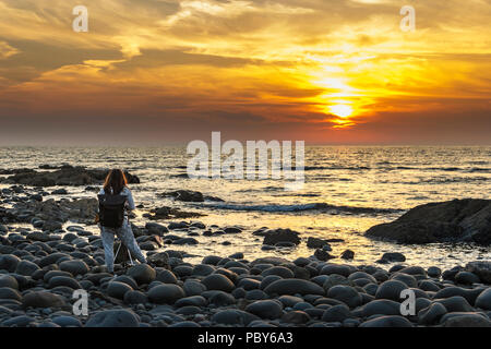 Une femme photographe capturant le spectaculaire coucher de soleil sur une plage de rochers à Westward Ho !, Devon, UK Banque D'Images