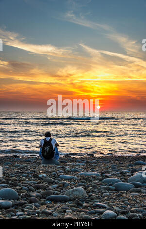 Un homme assis sur une plage de galets à regarder le coucher de soleil spectaculaire à Westward Ho !, Devon, UK Banque D'Images