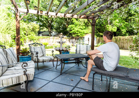 Jeune homme assis sur la chaise de salon patio extérieur en jardin de fleurs de printemps dans la cour porche d'accueil avec fontaine zen, pergola tonnelle auvent, table, pla Banque D'Images