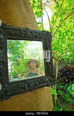 Le reflet d'une femme portant un chapeau de soleil et en robe d'un miroir suspendu à un arbre à l'extérieur en milieu tropical Banque D'Images