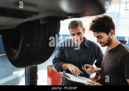 En uniforme de la mécanique des notes tout en se tenant par une voiture dans une station-service de l'automobile. Deux personnes travaillant dans un garage automobile et faire une liste des problèmes dans Banque D'Images