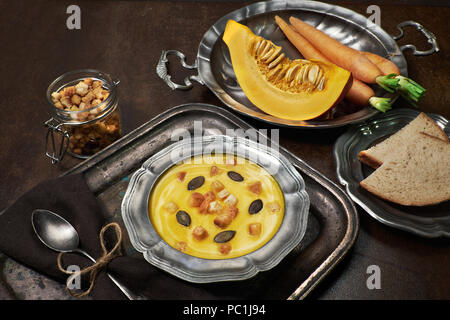 Crème de carotte et de potiron savoureux servi avec des croûtons dans la soupe en étain métal bol. Piieces citrouille sculptée, tas de carottes et des tranches de pain et sont aroun Banque D'Images