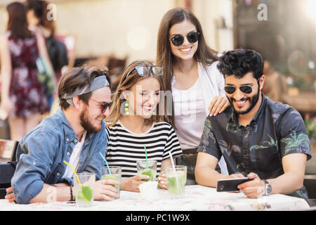 Portrait of cheerful young friends looking at smartphone while sitting in cafe, mixed race des gens assis à une table de restaurant en utilisant le téléphone mobile. Banque D'Images