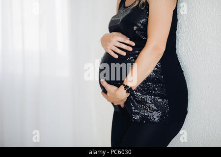 Close-up of méconnaissable pregnant woman in black élégant haut mousseux embrassant son ventre avec deux bras sur fond blanc. Copyspace. Banque D'Images