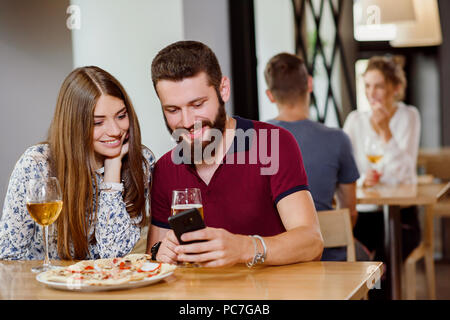 Sweet couple sitting, manger de la pizza, boire le vin blanc et la bière. Beau barbu et belle jeune fille à la recherche de téléphone et de sourire. L'intérieur de la pizzeria. Banque D'Images