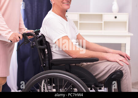 L'homme en fauteuil roulant qui est poussé par le fournisseur de soins. Image coupée de l'homme handicapé en fauteuil roulant poussé par l'infirmière. Arrière-plan de la salle de séjour. Banque D'Images