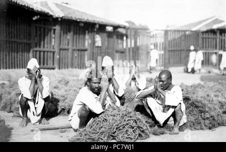 La photographie montre cinq hommes prisonniers accroupis avec mains pressées ensemble au milieu d'un matériau fibreux à Rangoon, Birmanie 1910-1920 Banque D'Images