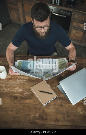 Beau jeune homme à lunettes lisant le journal tout en étant assis à table en bois Banque D'Images