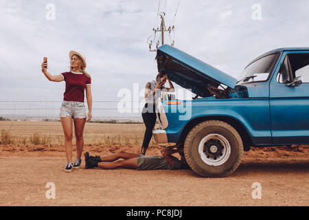 Les femmes qui essaient d'établir une communication pour l'assistance aux ventilées voiture. Une femme couché sous la voiture pour résoudre le problème on country road. Trois jeunes fem Banque D'Images