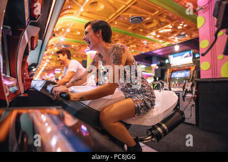 Couple heureux d'avoir l'amusement jouer des jeux de course arcade lors d'un salon de jeux. Excitée femme jouant un jeu de course arcade assis sur un vélo de course. Banque D'Images