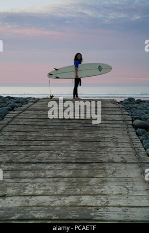 Surfeur femme debout sur la crête de galets avec surf avant de partir à la mer pour naviguer à Westward Ho ! Beach, North Devon, Angleterre, Europe. Banque D'Images