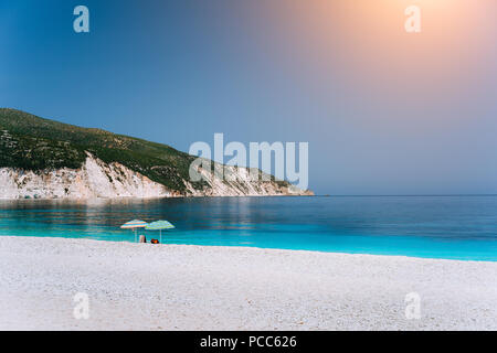 Soleil parasols sur une plage de galets avec une mer calme bleu azur, roches blanches et ciel clair en arrière-plan. Banque D'Images