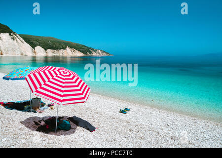 Soleil parasols sur une plage de galets avec une mer calme bleu azur, roches blanches et ciel clair en arrière-plan. Banque D'Images