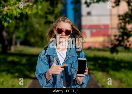 Lycéenne fille adolescente. L'été dans la nature. Lunettes de soleil en forme de coeur. Dans ses mains est titulaire d'un smartphone. Le concept de nouveau téléphone, d'une nouvelle demande. Espace libre pour le texte. Banque D'Images