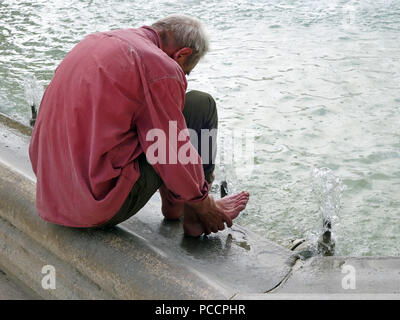 Vieil homme avoir chaud et inconfortable dans la canicule tente de se rafraîchir en mettant ses pieds dans la fraîcheur de l'eau de la fontaine Banque D'Images