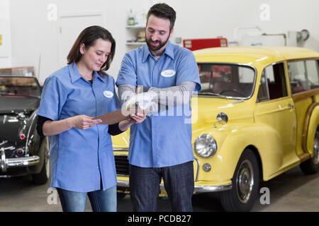 Une paire de mâles et femelles de race blanche parlons mécanique auto sur un bordereau de réparation dans un atelier de réparation de voitures classiques. Banque D'Images